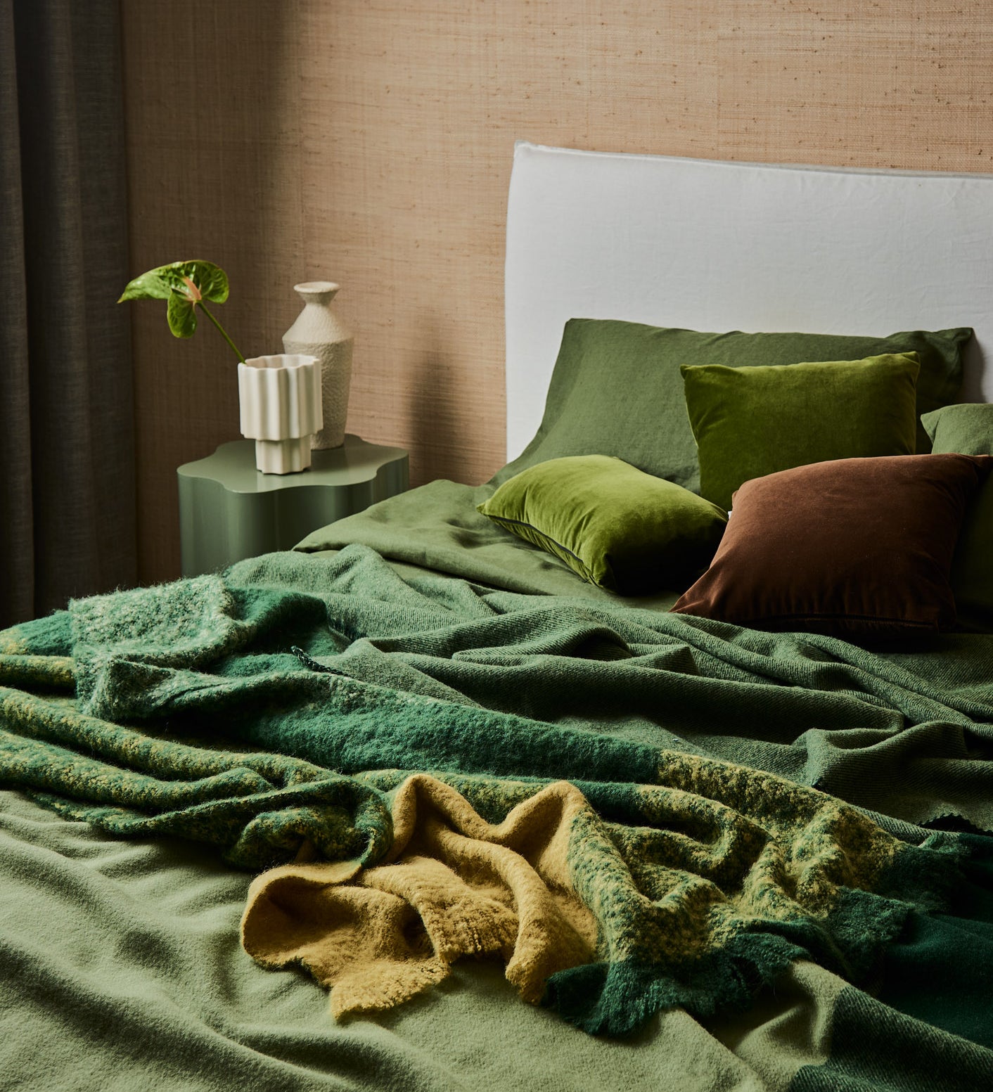 A rumpled bed made up with a Geo blanket in forest, draped with a Lustre throw in forest.