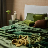 A tousled bed made up with the geo blanket in forest and draped with a lustre throw in forest.